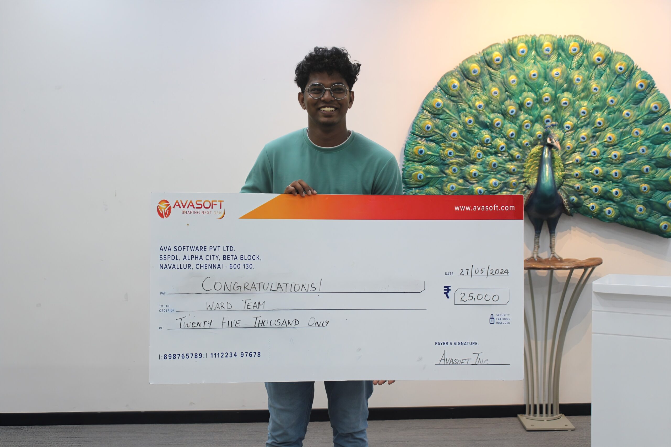 A man is smiling and holding a large check with the amount of ₹250,000, made out to a "Ward Team". The check is from AvaSoft. In the background, there is a decorative peacock artwork displayed on a wall.