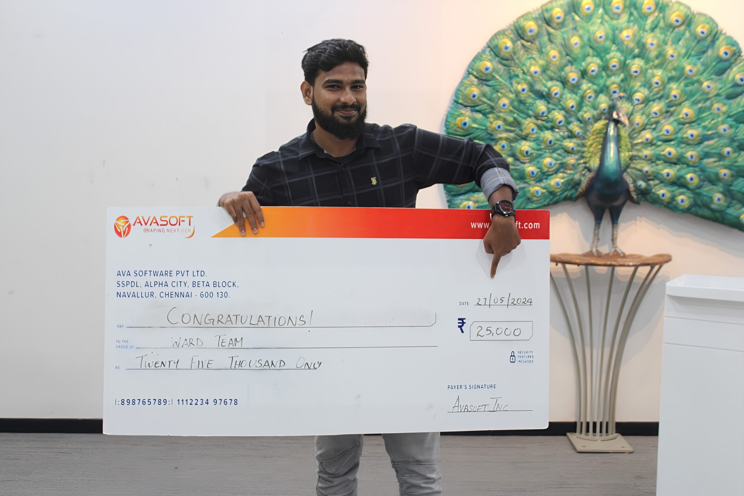 A man is smiling and holding a large check with the amount of ₹250,000, made out to a "Ward Team". The check is from AvaSoft. In the background, there is a decorative peacock artwork displayed on a wall.