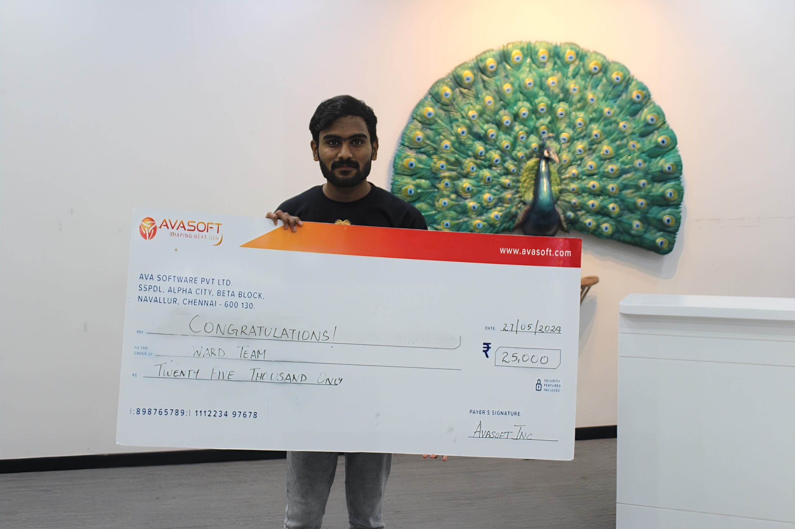 A man is smiling and holding a large check with the amount of ₹250,000, made out to a "Ward Team". The check is from AvaSoft. In the background, there is a decorative peacock artwork displayed on a wall.