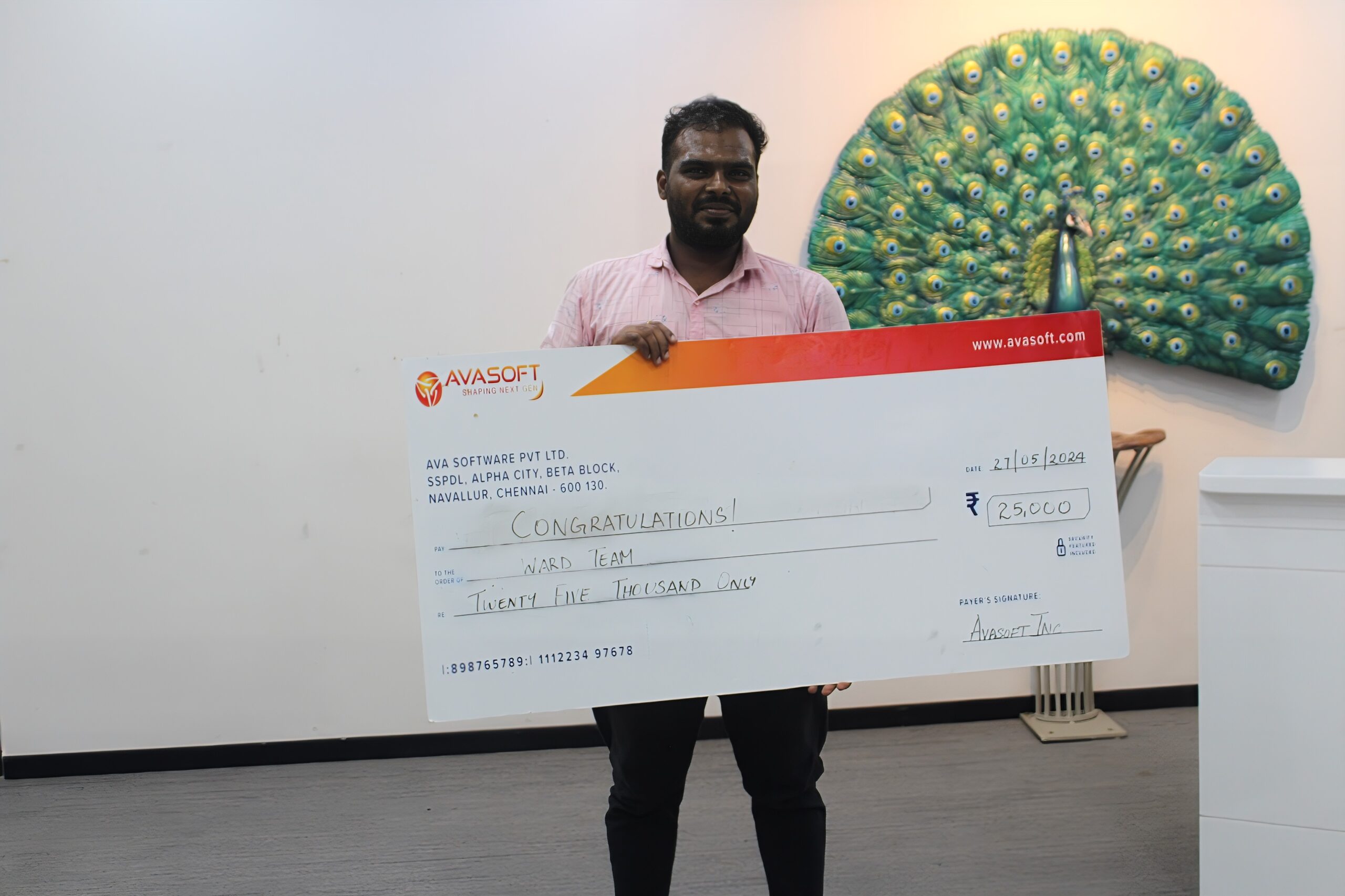 A man is smiling and holding a large check with the amount of ₹250,000, made out to a "Ward Team". The check is from AvaSoft. In the background, there is a decorative peacock artwork displayed on a wall.