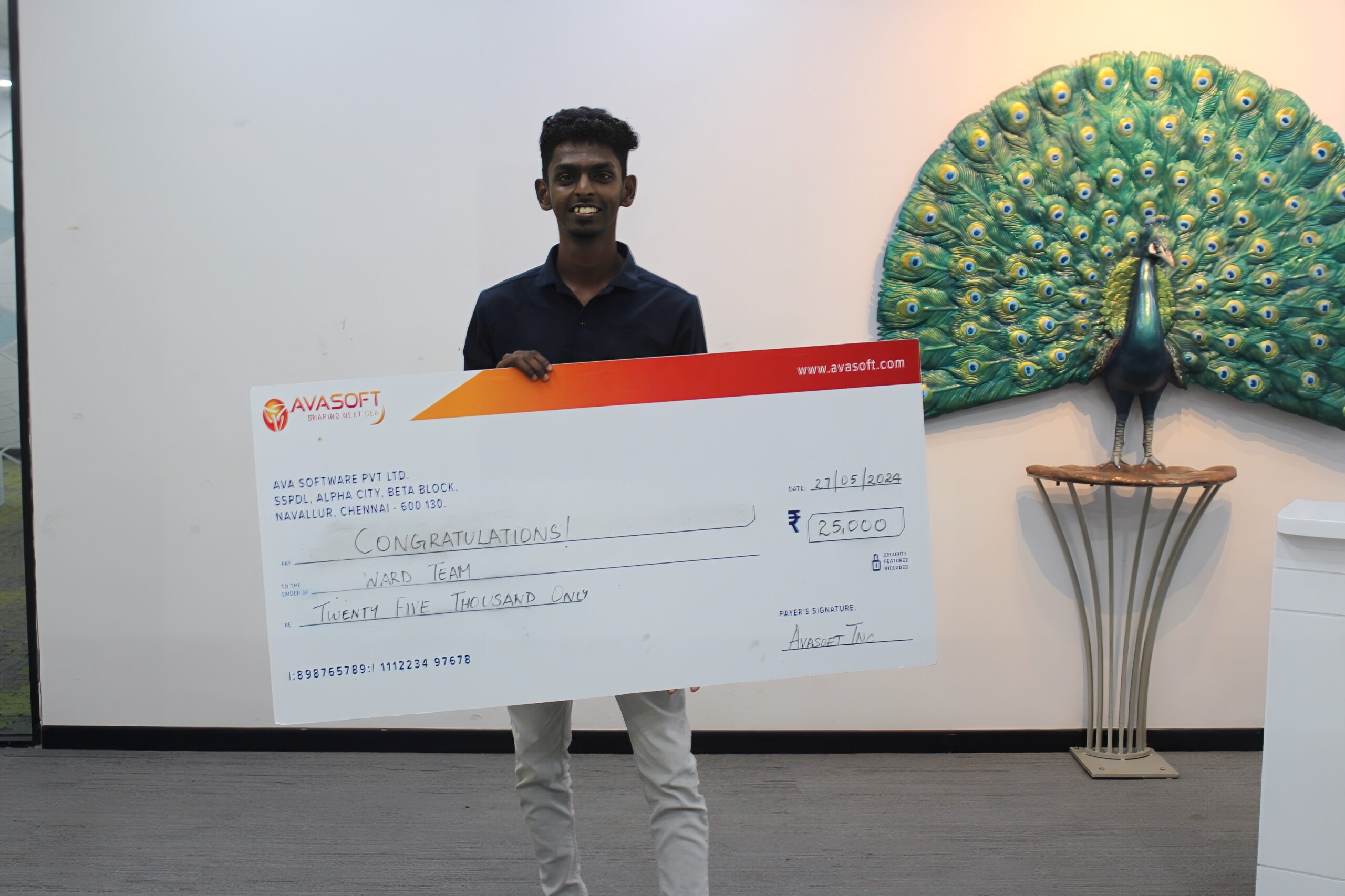 A man is smiling and holding a large check with the amount of ₹250,000, made out to a "Ward Team". The check is from AvaSoft. In the background, there is a decorative peacock artwork displayed on a wall.